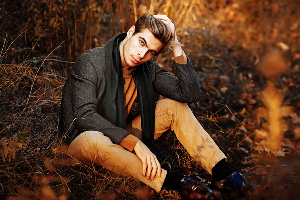 a man, a model in a gray jacket, a green scarf and beige pants and a sweater sits on the grass in autumn, among the autumn trees. posing. thoughtfulness and sadness. - Foto, imagen