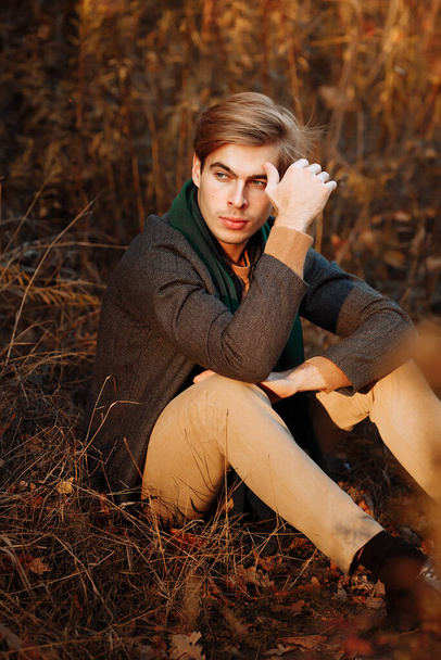 un uomo, un modello in giacca grigia, una sciarpa verde e pantaloni beige e un maglione siede sull'erba in autunno, tra gli alberi autunnali. In posa. pensosità e tristezza. - Foto, immagini
