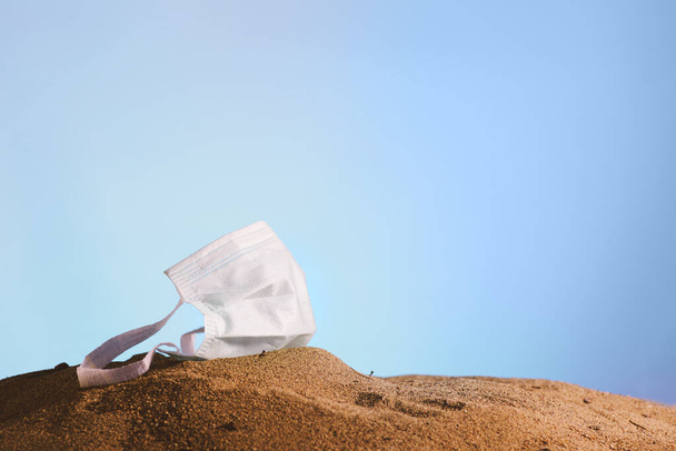Medical mask tossed on sand and on a blue sky background. - 写真・画像