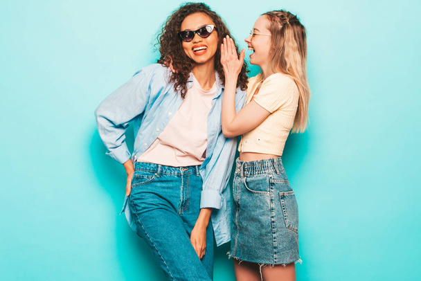 Two young beautiful smiling international hipster female in trendy summer clothes. Sexy carefree women posing near blue wall in studio. Positive models having fun. Tells her friend secret - 写真・画像