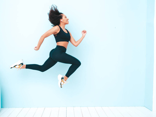 Fitness smiling black woman in sports clothing with afro curls hairstyle.She wearing sportswear. Young beautiful model with perfect tanned body.Female jumping and running in studio near blue wall - Foto, imagen