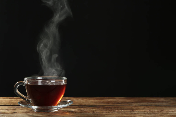 Cup with steam on wooden table against black background. Space for text - Fotografie, Obrázek