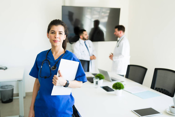 Tired caucasian nurse in blue scrubs making eye contact with a serious expression while holding the medical chart with the information of a patient - Photo, Image