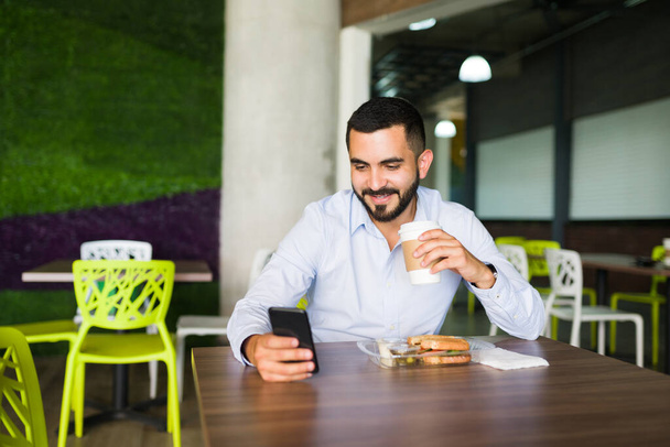 Jeune homme attrayant buvant du café et mangeant un sandwich à la cafétéria du travail pendant sa pause déjeuner en regardant les médias sociaux et en envoyant des SMS sur son smartphone - Photo, image