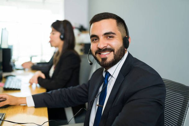 Egy latin fiatalember arcképe öltönyben és fülhallgatóval az asztalánál egy call centerben. Üzleti képviselő, aki egy technológiai támogató cégnél dolgozik.  - Fotó, kép