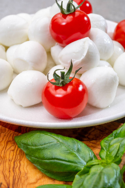 Włoski tricolore, małe kulki świeżego białego miękkiego włoskiego sera mozzarella, dojrzałe czerwone pomidory wiśniowe i świeże zielone zioła bazylii, gotowe do robienia sałatki caprese, zbliżenie - Zdjęcie, obraz