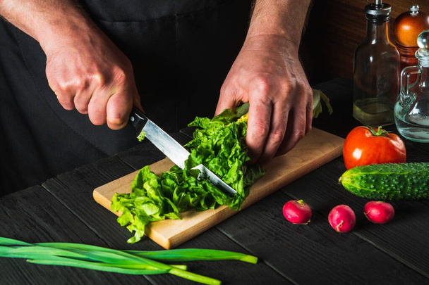 Chef or cook cuts fresh napa cabbage with a knife for salad on a vintage kitchen table with fresh vegetables. Cooking and restaurant or cafe concept - Photo, Image