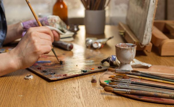 The artist is working on a Still Life. With a brush, he mixes oil paints on the palette.Studio with a desktop easel and brushes on the table. Close-up,Focus on the bunch of brushes in the foreground  - Photo, Image