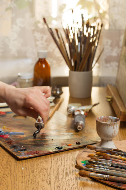 Las manos de una artista femenina exprimen pintura marrón de un tubo viejo. Artistas lugar de trabajo, enfoque selectivo, marco vertical.Enfoque en el montón de pinceles en primer plano - Foto, Imagen