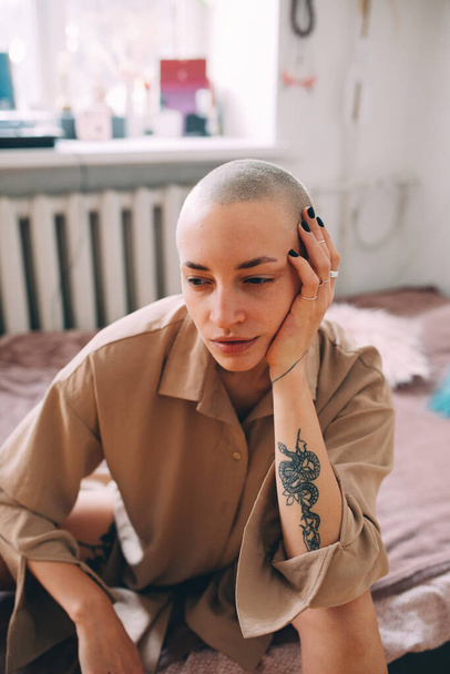 Girl with short haircut and black tattoos sitting at the bed and looking away - Valokuva, kuva