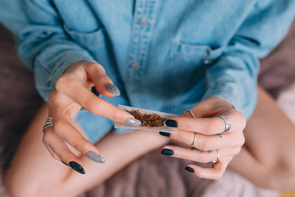 Woman wearing jeans shirt making hand rolled cigarette with tobacco at home - Foto, Imagem