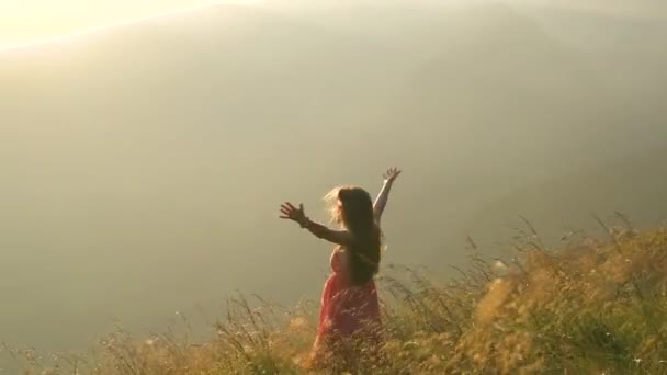 Vista posteriore di una donna felice escursionista in abito rosso in piedi su una collina erbosa in una serata ventosa in montagna autunnale con braccia aperte godendo della vista della natura al tramonto. - Filmati, video