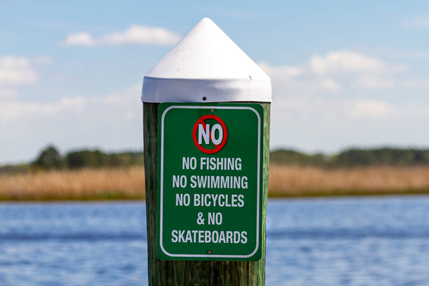Ein Schild an einem Steg am Meer weist darauf hin, dass Schwimmen, Tauchen, Radfahren oder Skateboarden auf der Seebrücke verboten ist. Vielseitiges Image mit Gesundheits- und Sicherheitsaspekten oder restriktiven Vorschriften, die Freiheitskonzepte einschränken.  - Foto, Bild