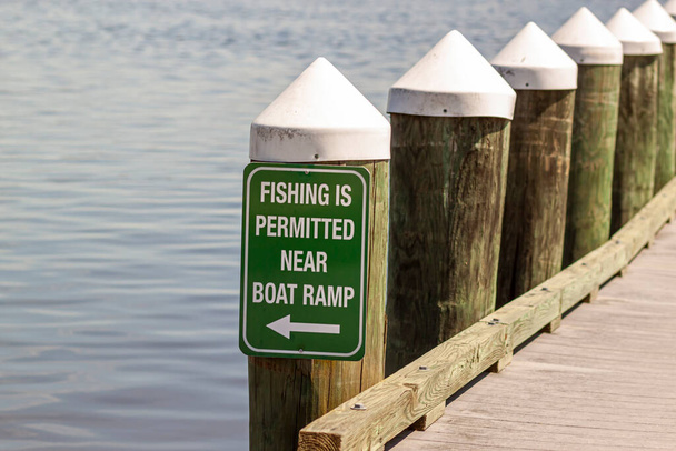 Fishing is permitted near boat ramp sign on green metal plate attached to a dock post on the waterfront of a local harbor. - Photo, Image