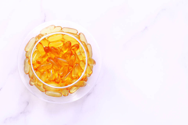 Dietary supplement omega capsules in a round glass vase, yellow transparent vitamins on a marble table, healthy lifestyle and health care concept - Photo, Image