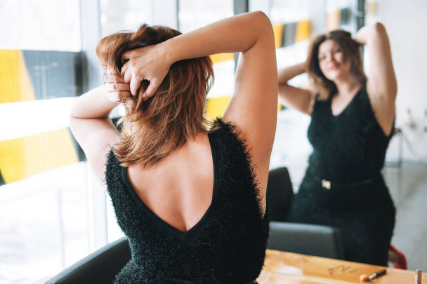 Schöne lächelnde junge Frau plus Größe Körper positiv in schwarzen Abendkleid Make Frisur in der Nähe Spiegel im Schönheitsstudio - Foto, Bild