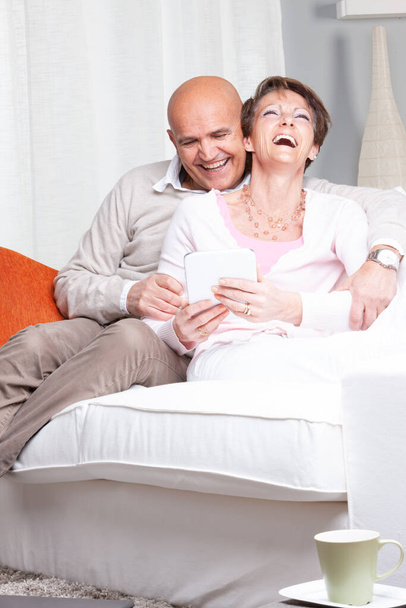 Fun loving married middle-aged couple enjoying a good joke laughing merrily as they relax on a sofa sharing media on a tablet-pc - 写真・画像