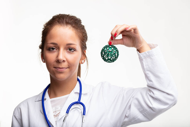 Young medical doctor or nurse holding up a decorative green Christmas bauble in her hand with a friendly smile to celebrate the holiday season on white - Foto, afbeelding