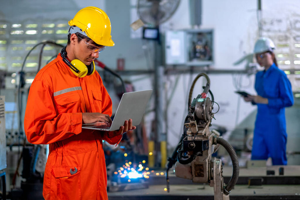 Factory worker man hold laptop to monitor metal welding robotic machine and co-worker in background of workplace area. Industrial and technology support system help in working of employee concept. - Foto, imagen
