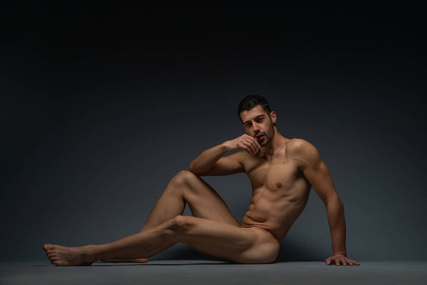 Gorgeous naked male model sitting isolated on the floor in a studio while looking in the camera - Foto, afbeelding