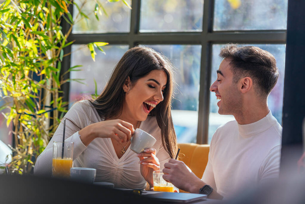 Two happy young people are laughing while sitting at a table in indoor cafe - Foto, Imagen