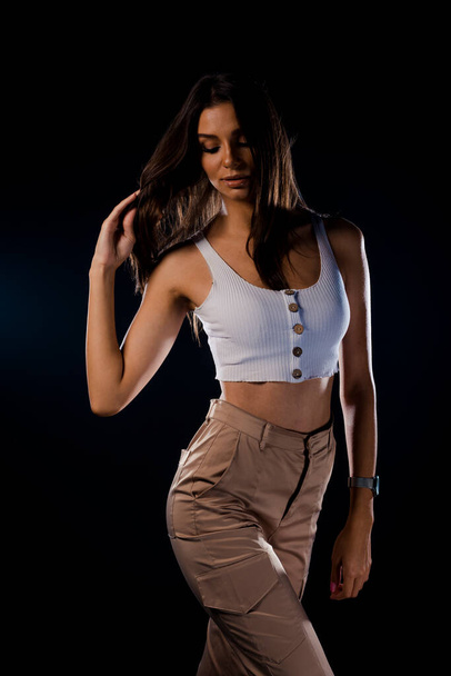 Молодая сексуальная стильная женщина в летней модной одежде в стильных брюках стоит изолированно на темной стене - Фото, изображение