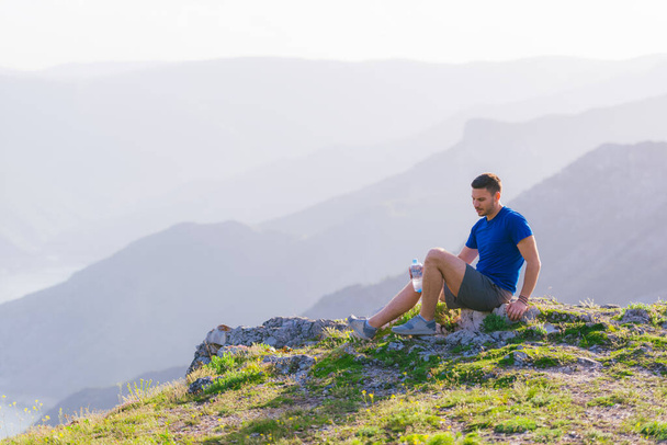 Schöner Athlet sitzt auf einem felsigen Gipfel und blickt auf die atemberaubende Bergkette und einen wunderschönen See, während er ein blaues Hemd und graue Shorts trägt. - Foto, Bild