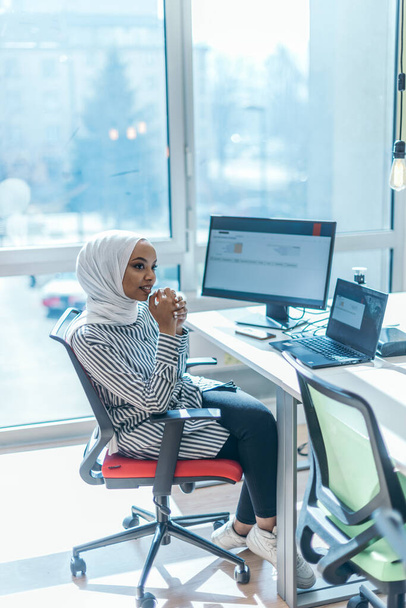 Wirtschaftspädagogik und Bürokonzept. Afroamerikanische Geschäftsfrau trägt einen Hidschab und tippt auf einem PC (Computer). Unternehmerin sitzt auf ihrem Schreibtisch. - Foto, Bild