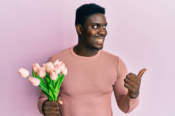 Yakışıklı, elinde pembe lale buketi olan siyah bir adam. Çiçekler başparmağıyla yan tarafa bakıyor. Gülümsüyor.  - Fotoğraf, Görsel