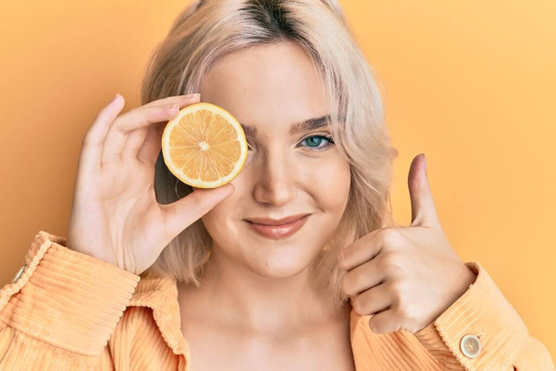 Jong blond meisje met citroen over het oog glimlachend gelukkig en positief, duim omhoog doen uitstekend en goedkeuring teken  - Foto, afbeelding