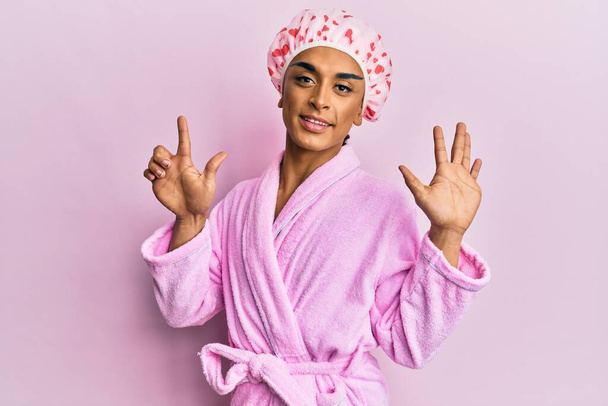 Spaanse man draagt make-up met een badmuts en badjas en wijst naar boven met vingers nummer zeven en glimlacht zelfverzekerd en gelukkig.  - Foto, afbeelding