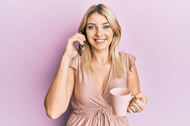 Νεαρή καυκάσια γυναίκα που μιλάει στο smartphone και πίνει ένα φλιτζάνι καφέ χαμογελώντας με ένα χαρούμενο και δροσερό χαμόγελο στο πρόσωπο. που δείχνει δόντια.  - Φωτογραφία, εικόνα