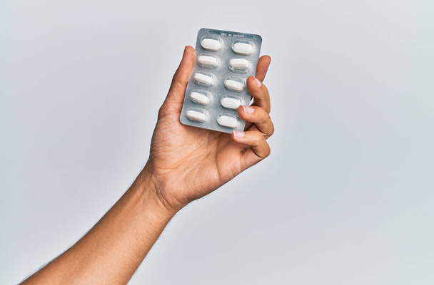 Hand of hispanic man holding pills over isolated white background. - Foto, afbeelding