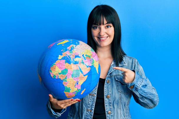Joven mujer hispana sosteniendo la bola del mundo sonriendo feliz señalando con la mano y el dedo  - Foto, Imagen