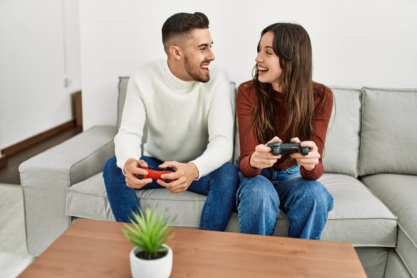 Νεαρό ζευγάρι ισπανόφωνων που παίζει βιντεοπαιχνίδι κάθεται στον καναπέ στο σπίτι. - Φωτογραφία, εικόνα