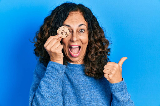 Středního věku hispánská žena drží sušenku přes oko ukazuje palec nahoru na stranu usmívá šťastný s otevřenými ústy  - Fotografie, Obrázek