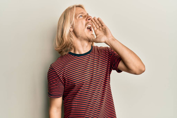 Homem jovem caucasiano com cabelos longos vestindo camisa listrada casual gritando e gritando alto para o lado com a mão na boca. conceito de comunicação.  - Foto, Imagem