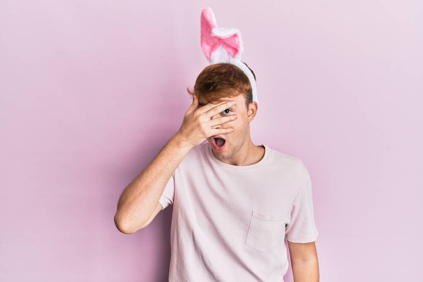 Giovane uomo caucasico indossa cute orecchie coniglietto pasquale sbirciare in shock che copre il viso e gli occhi con la mano, guardando attraverso le dita paura  - Foto, immagini