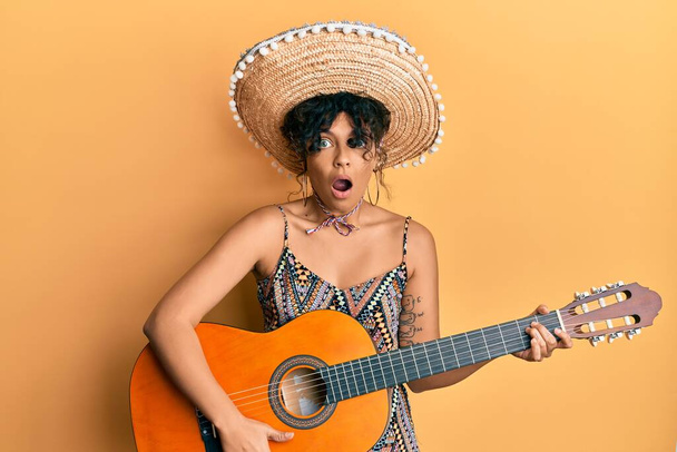 Jeune femme hispanique tenant un chapeau mexicain jouant de la guitare classique effrayée et choquée par la surprise et l'expression étonnante, la peur et le visage excité.  - Photo, image