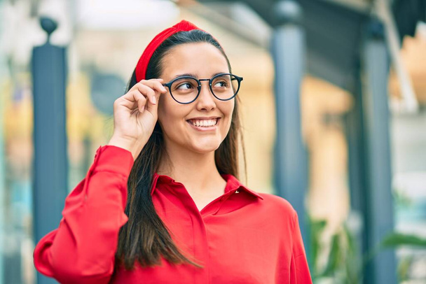Joven mujer hispana sonriendo feliz tocando sus gafas en la ciudad. - Foto, Imagen
