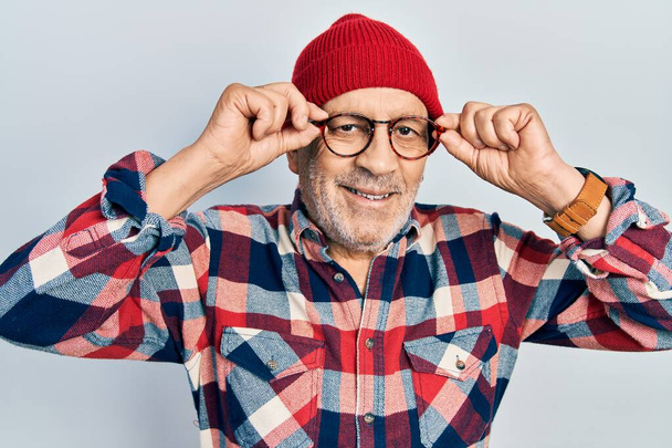 Bonito homem maduro segurando óculos, usando chapéu de lã sorrindo com um sorriso feliz e legal no rosto. mostrando dentes.  - Foto, Imagem