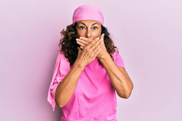 Middle age hispanic woman wearing breast cancer support pink scarf shocked covering mouth with hands for mistake. secret concept.  - Foto, Bild