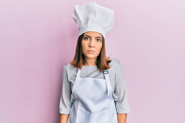 Mujer rubia joven con uniforme de cocinero profesional y sombrero enojado y loco levantando el puño frustrado y furioso mientras gritaba con ira. rabia y concepto agresivo.  - Foto, Imagen