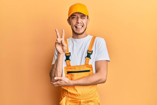 Hispanic young man wearing handyman uniform smiling with happy face winking at the camera doing victory sign with fingers. number two.  - Foto, afbeelding