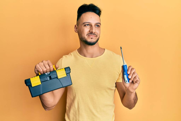 Hombre árabe joven sosteniendo caja de herramientas y destornillador sonriendo mirando hacia un lado y mirando hacia otro pensando.  - Foto, imagen