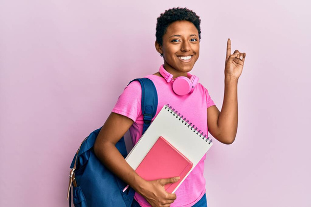 Молода афроамериканська дівчинка у шкільній сумці і тримає книжки здивовані ідеєю або питанням, що вказує пальцем на щасливе обличчя, по-перше.  - Фото, зображення