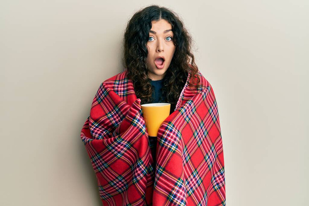 Young brunette woman with curly hair wrapped in a red warm red blanket drinking coffee afraid and shocked with surprise and amazed expression, fear and excited face.  - Photo, Image