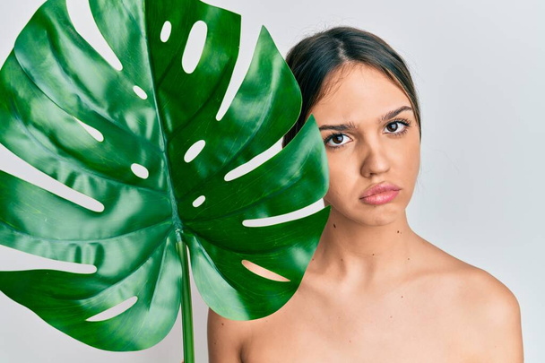 Young brunette woman holding green plant leaf close to beautiful face skeptic and nervous, frowning upset because of problem. negative person.  - Фото, изображение
