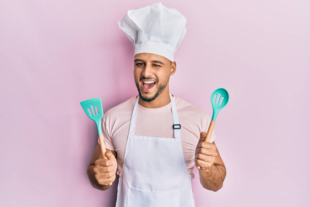 Mladý arabský muž na sobě profesionální kuchař zástěra a klobouk držící lžíci mrkající na fotoaparát se sexy výraz, veselý a šťastný obličej.  - Fotografie, Obrázek