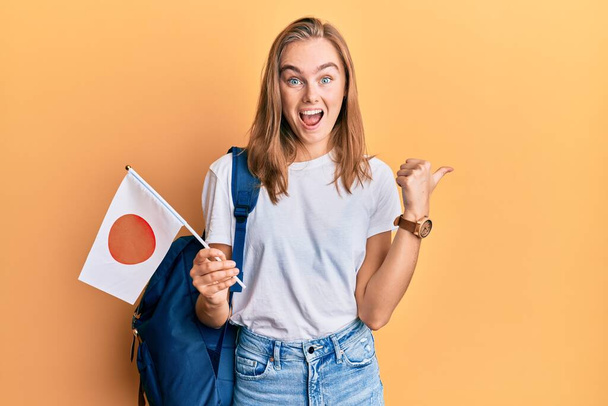 Güzel sarışın değişim öğrencisi. Elinde Japon bayrağı var. Parmağını kaldırmış, ağzı açık gülümsüyor.  - Fotoğraf, Görsel